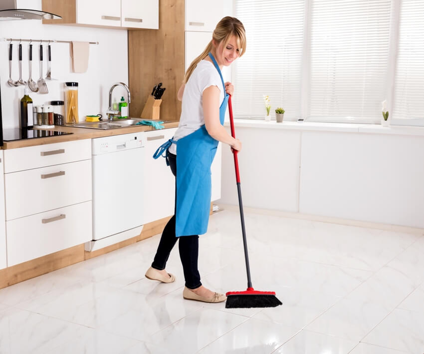 A woman with apron cleaning floor with a push broom in kitchen 