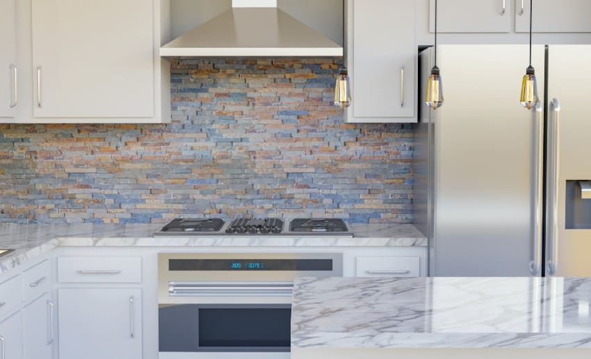A bright contemporary kitchen with white cabinets, marble counters, multicolor slate backsplash and stainless steel appliances with hanging lights above the island