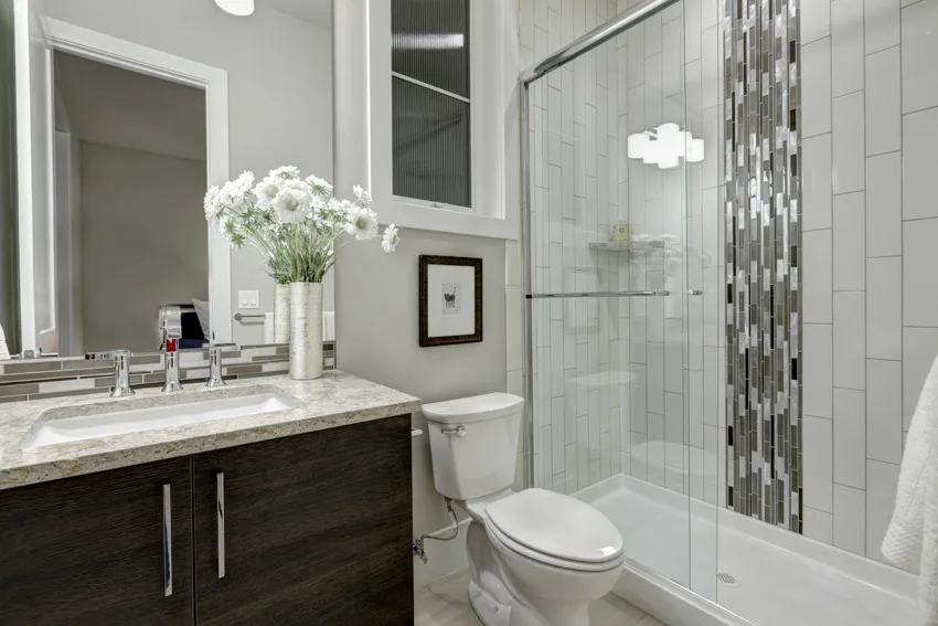 Bathroom with waterfall tile shower, toilet, glass door, countertop, mirror, cabinet, and sink