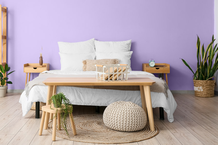 Bedroom with lavender painted wall, pillows, comforter, wood bench, ottoman, rug, indoor plant, and lavender painted wall