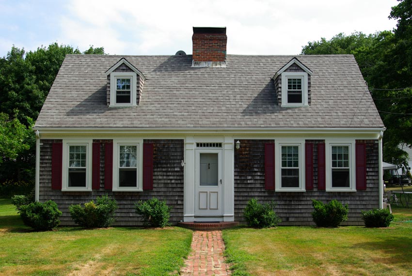 Cape cod house with historic colors, chimney, dormer, windows, front door, hedge plants, and landscaped lawn