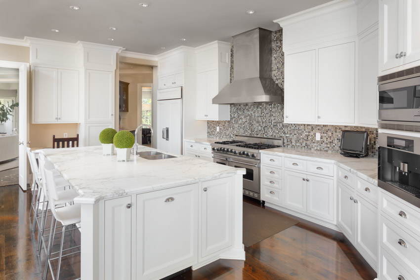 Kitchen with white cabinets, backsplash, countertop, island, high chairs, oven, stove, and range hood