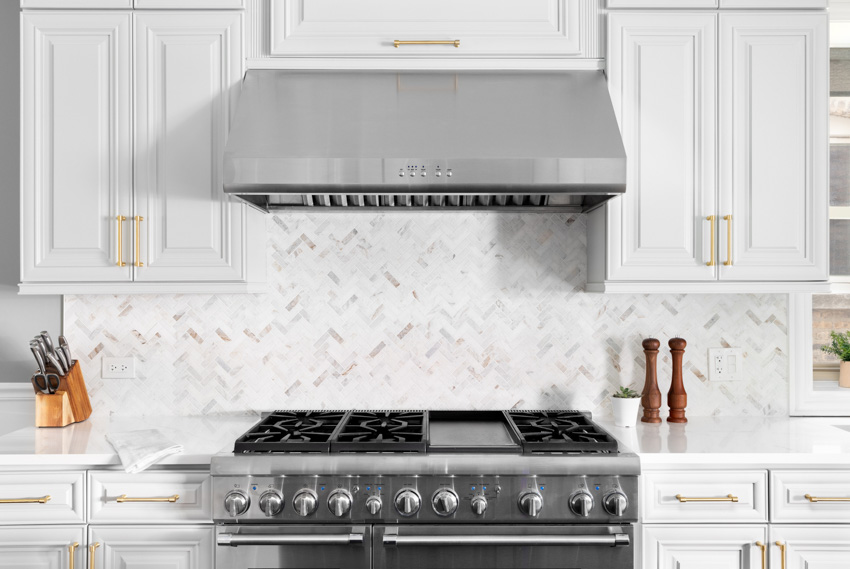 Contemporary kitchen with white cabinets, range hood, stove, countertop, and herringbone Calcatta gold backsplash