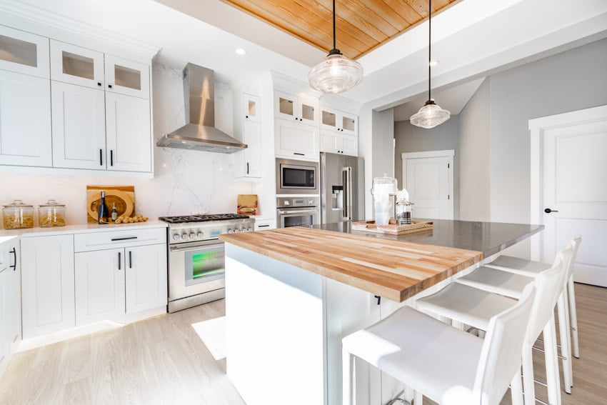 Farmhouse kitchen with white cabinets, chairs, pendant lights, range hood, oven, wood flooring, and Carrara marble slab backsplash