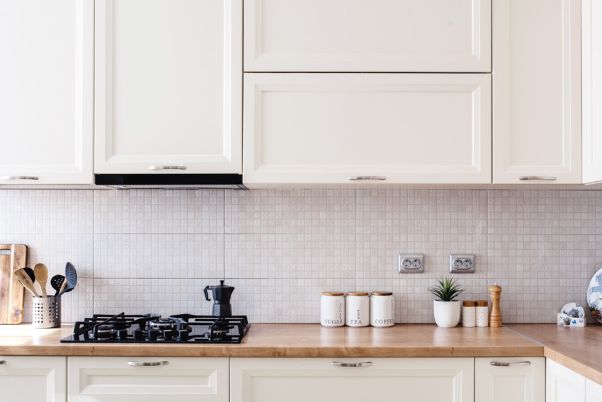 Farmhouse kitchen with wood countertop, ceramic mosaic tile backsplash, white cabinets, stove, and range hood