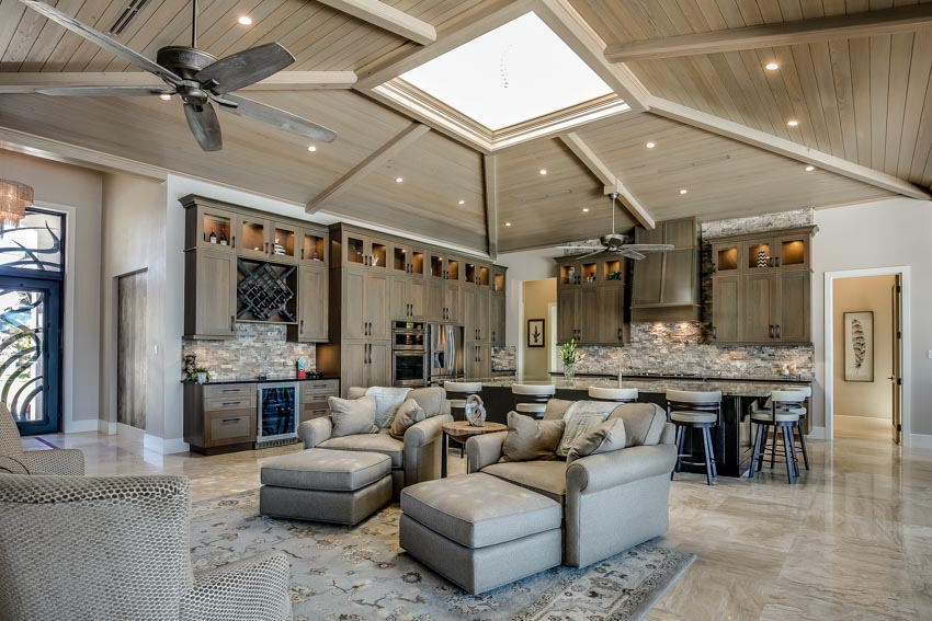 Farmhouse living room with skylight window, beadboard ceiling, cushioned chairs, ottomans, kitchen space, cabinets, backsplash, and ceiling fan