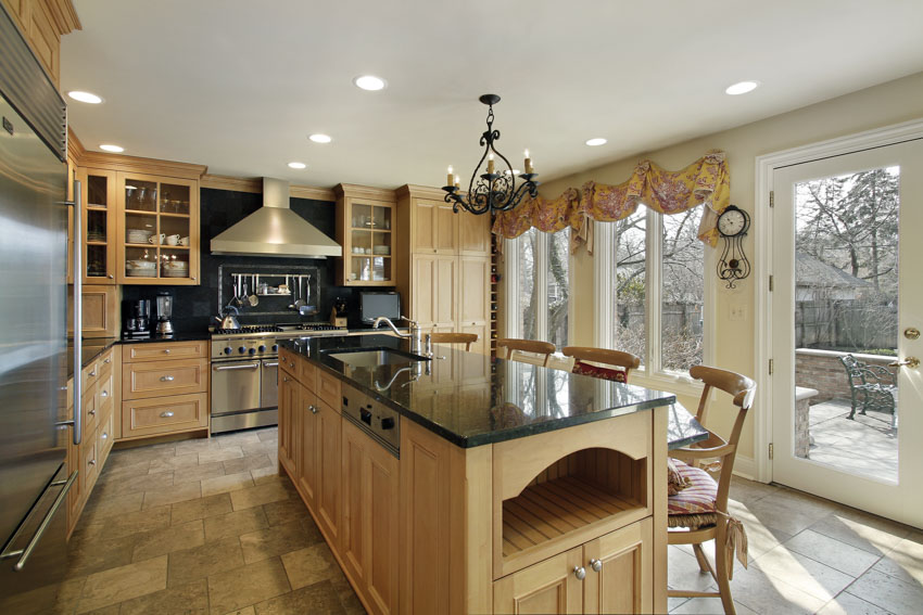 Kitchen with black countertops, oak cabinets, island, tile flooring, range hood, backsplash, chandelier, glass door, and windows