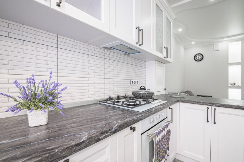 Kitchen with black fantasy quartzite countertop, tile backsplash, stove, oven, and white cabinets