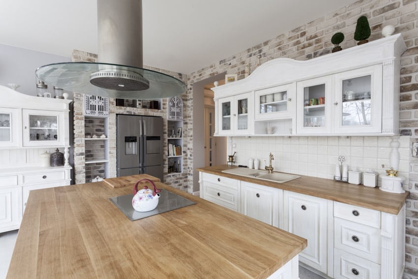 Kitchen with brick walls, stove, island, range hood, glass insert cabinets, refrigerator, backsplash, sink, and faucet