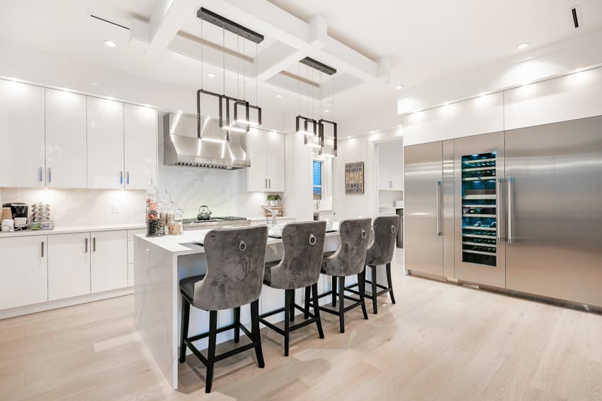 Kitchen with lighting above modern white lacquer cabinets, silver hardware, quartz waterfall, island, and barrel back bar stools