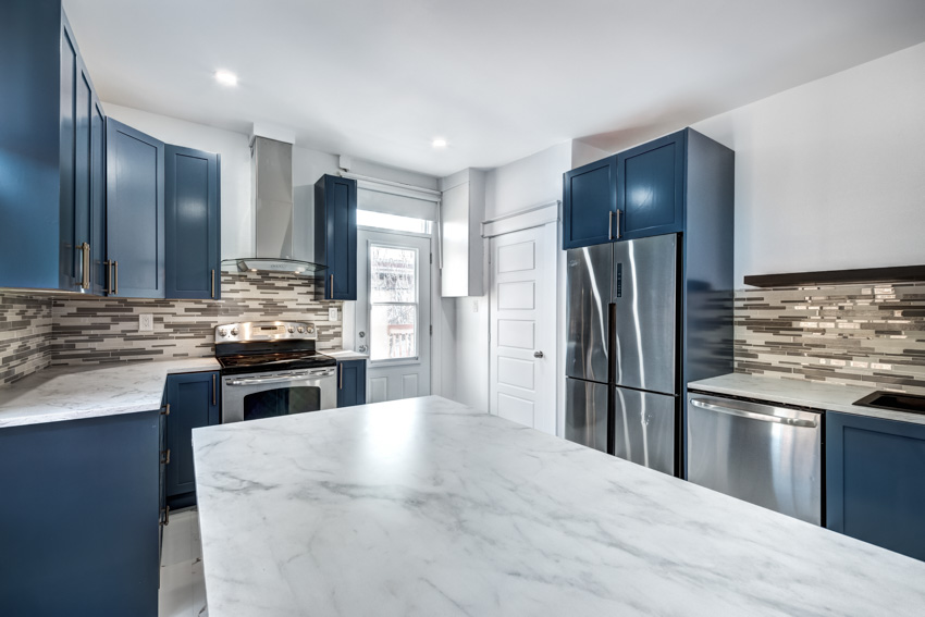 Modern kitchen with blue cabinets, honed Carrara marble countertop, island, refrigerator, and tile backsplash