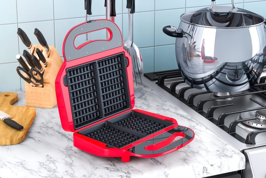 Opened red waffle iron on the marble kitchen countertop with cooktop on the side
