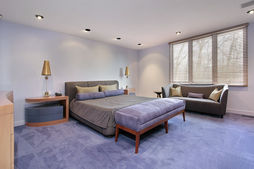 Simple bedroom with nightstand, bench seating, couch, carpet flooring, lavender painted wall, lamp, and windows