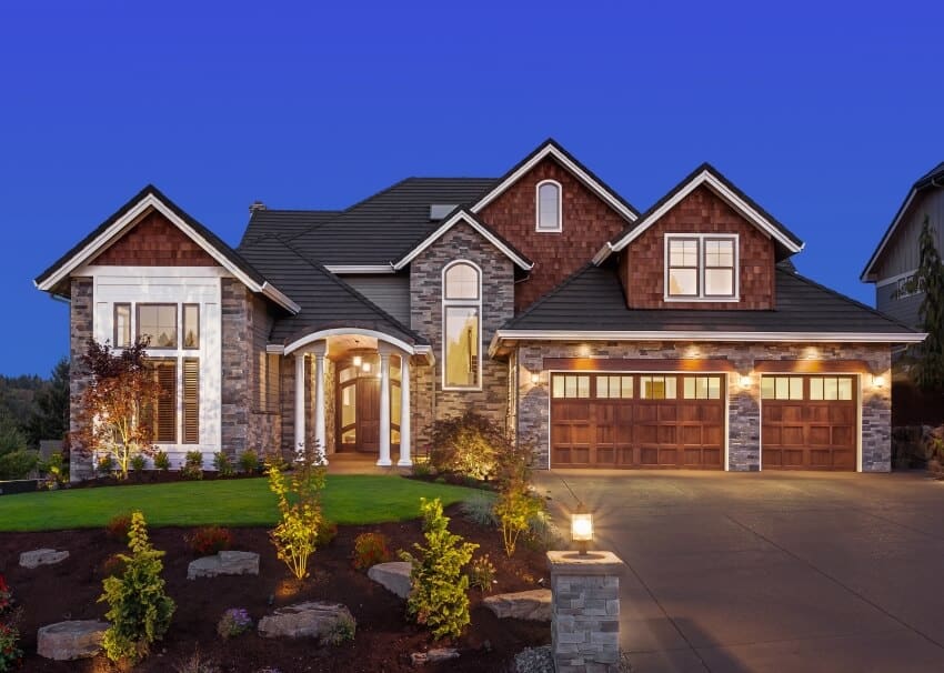 Stone exterior of a luxurious house with black roof, porch columns, and wooden garage doors