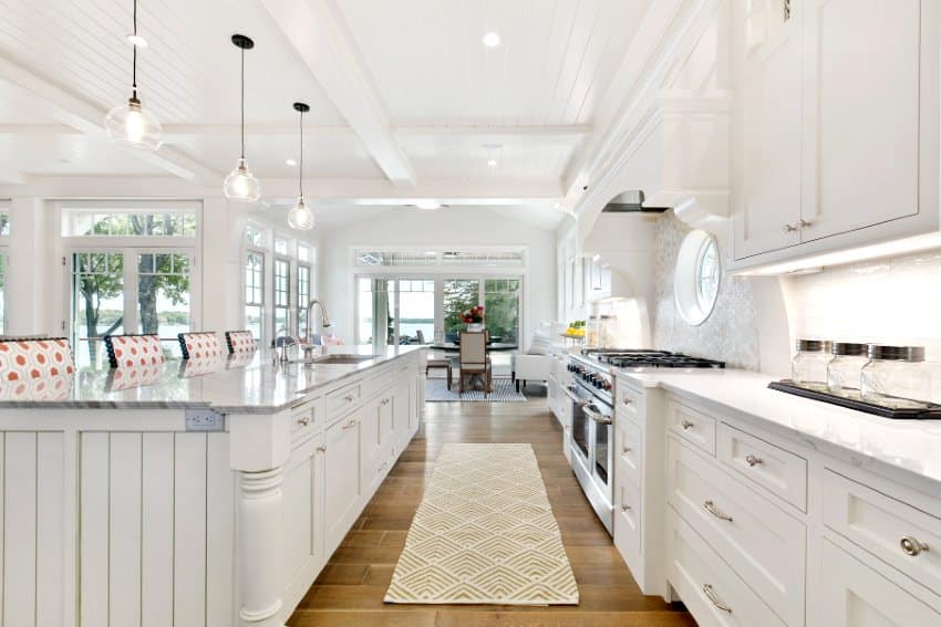 White kitchen galley of new home with wooden floors, vertical shiplap kitchen island, white cabinets and beautiful port window behind the cooktop
