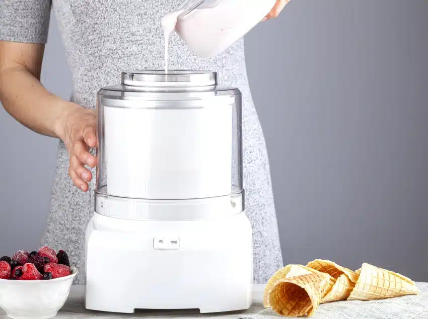 Woman is pouring a mixture into an ice cream maker with mixed berries and waffle cones on the side