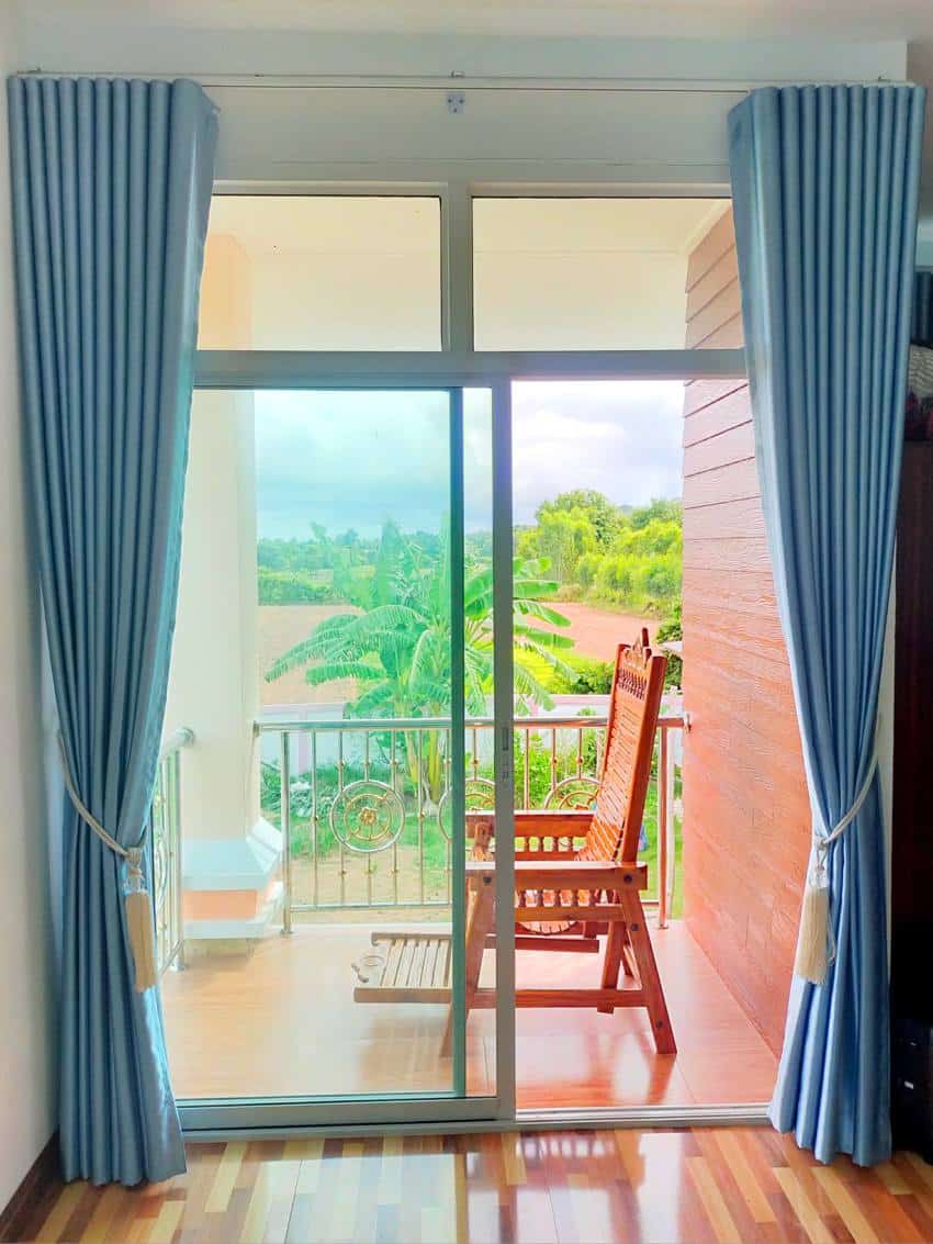 A room with blue portiere curtains hanged above white glass doors connected to a balcony with wooden armchair and outside view of a green field