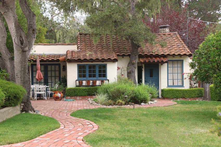 Cottage suburban house with brick tile walkway, orange shingle roof, chimney, windows, outdoor patio, front door, and hedge plants