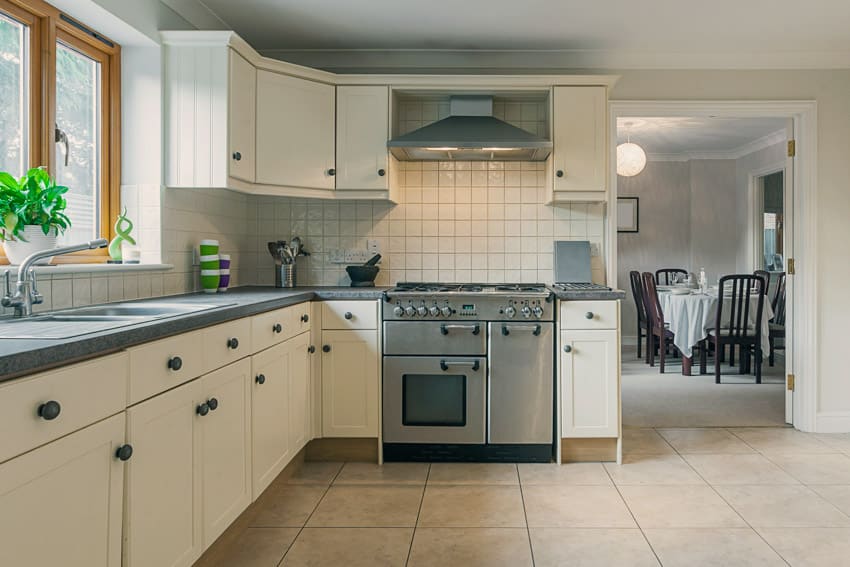 Kitchen with white square tile backsplash, oven, cabinets, stove, range hood, tile floors, and window