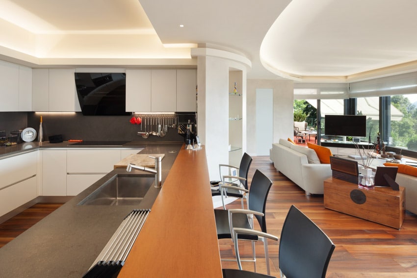 Modern kitchen next to living room with bar counter, cove ceiling lighting, white cabinets, backsplash, and countertop