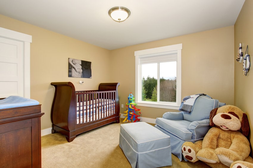 Nursery with crib, cushioned chair, pouf, window, and ceiling light