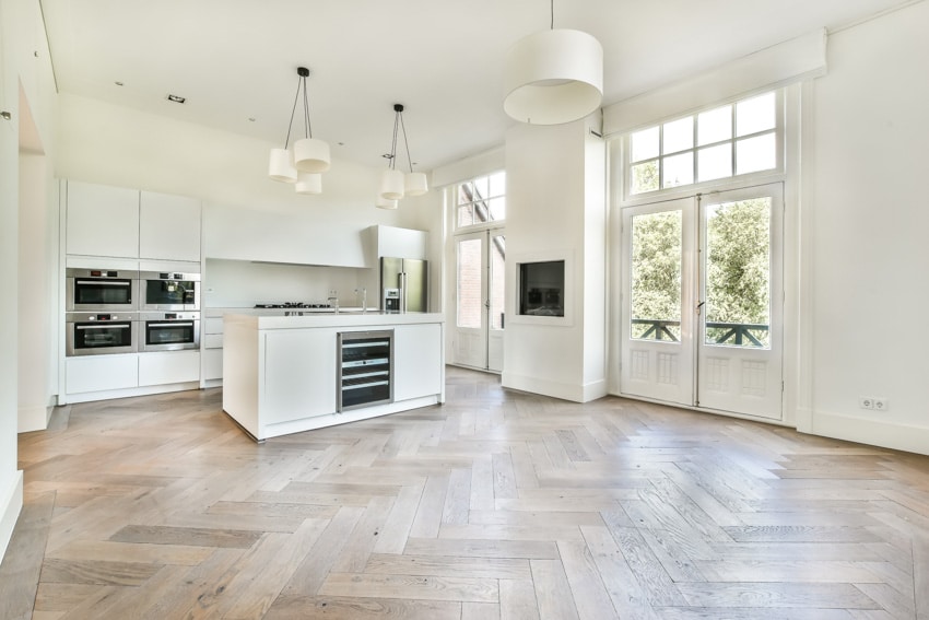 Remodeled minimalist kitchen with wood herringbone floor, island, white cabinets, oven, pendant lights, and glass door