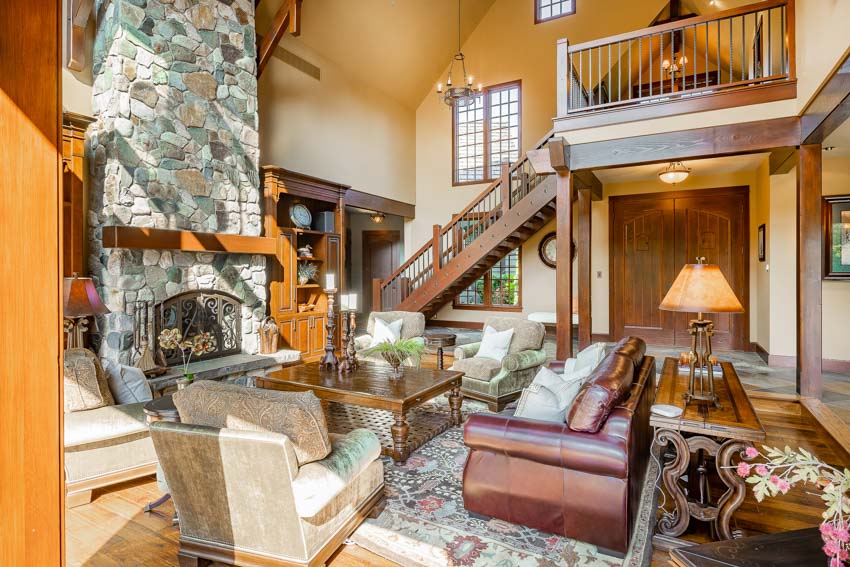 Rustic living room with cream paint colors, stone fireplace, sofa, chairs, coffee table, sofa, table, and staircase