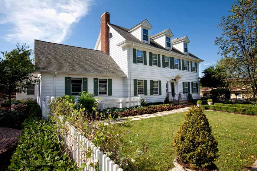 Traditional house exterior design with faux chimney, pitched roof, dormers, white siding, windows, picket fence, and lawn