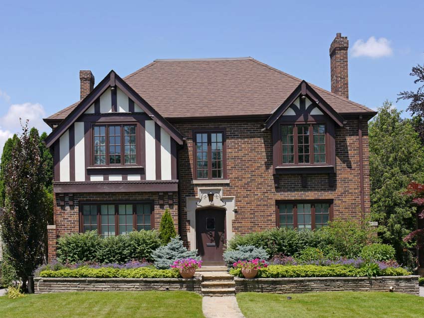 Tudor suburban house with brick wall cladding, windows, chimneys, front door, hedge plants, and lawn