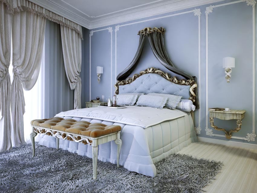 Classic light blue bedroom with rug, bench, headboard, bedding, headboard, pillows, wall sconces, window, and curtains