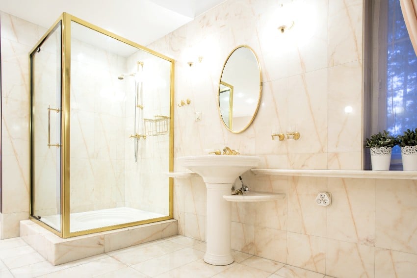 Classy bathroom in white and gold tones features marble floors and walls, glass shower and pedestal sink