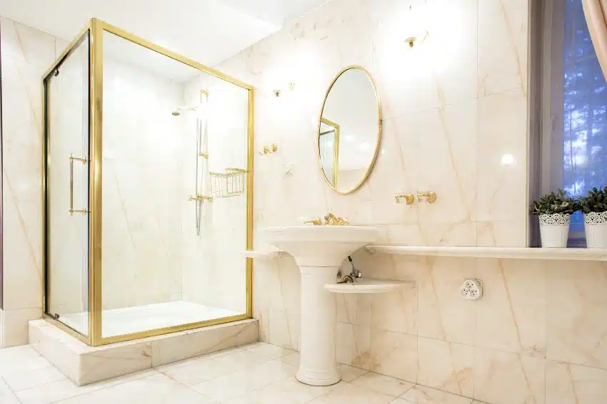 Classy bathroom in white and gold tones features marble floors and walls, glass shower and pedestal sink