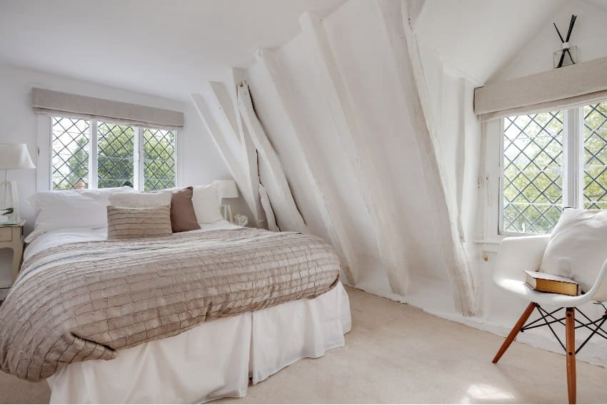 Cottage bedroom decorated in a light modern style with neutral color including bed with quilt and cushions leaded windows and numerous roof timbers