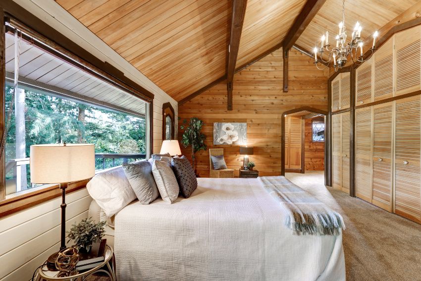 Cottage bedroom style interior with wood ceiling and beams, grey carpet floor, large bed, window and floor to ceiling wardrobe