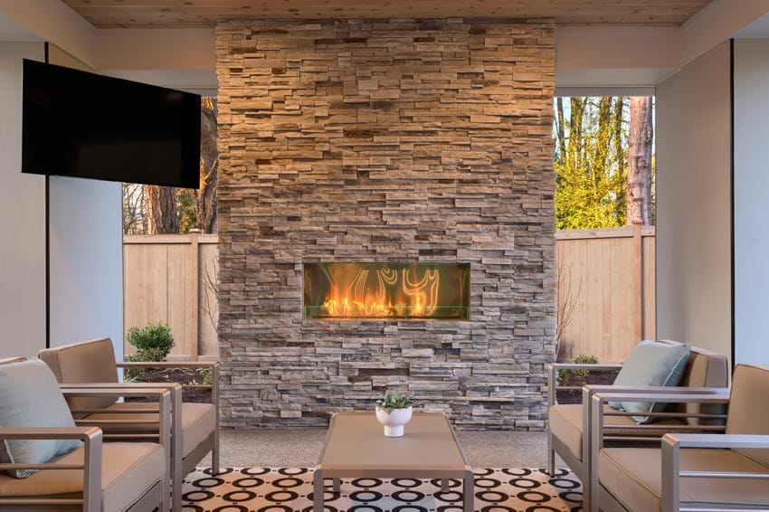Covered outdoor patio with cushioned chairs, rug, television, and ledger stone fireplace