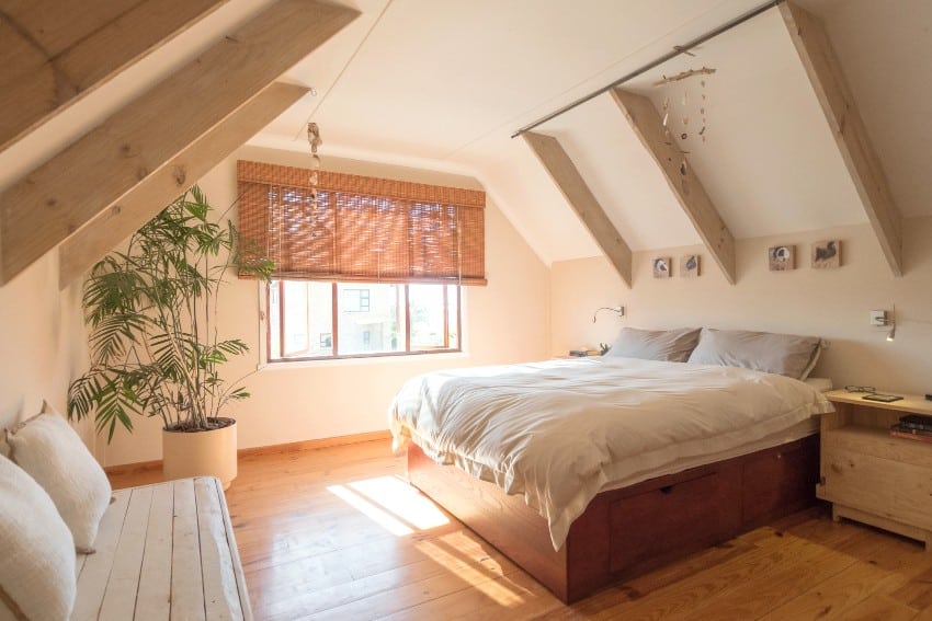 Cozy holiday cottage style bedroom with beige bedding, white ceiling with wooden beams, green indoor plant and small bench with cushions