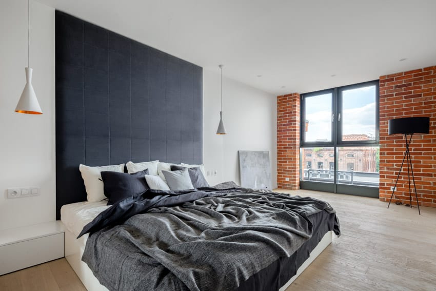 Cozy industrial bedroom with with black headboad, comforter, pillows, pendant lights, brick accent wall, floor lamp, and window