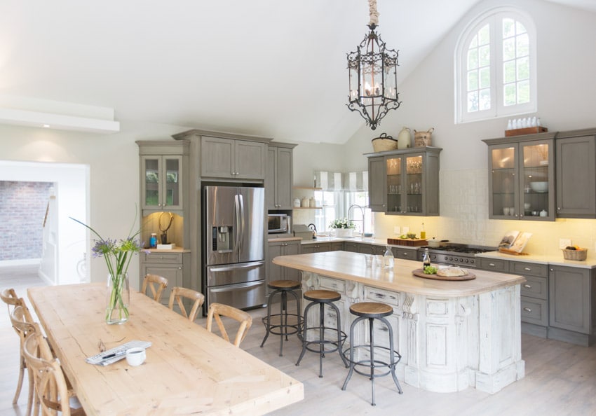 Farmhouse kitchen with bronze chandelier, bar stools, gray cabinets, and rustic whitewashed island,