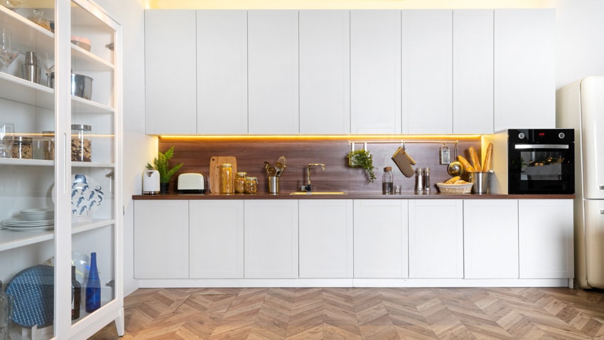 Farmhouse kitchen with white laminate cabinets, wood backsplash, glass cabinet, and wooden herringbone floors