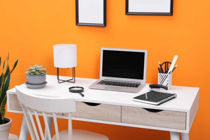 Home office with laptop stationery on desk and chair near orange wall