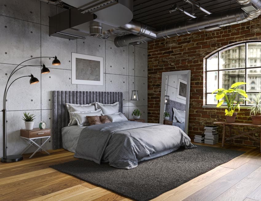 Industrial bedroom with pipes, bedding, rug, wood floor, concrete panel wall, floor lamp, nightstand, headboard, brick accent wall, mirror, and window