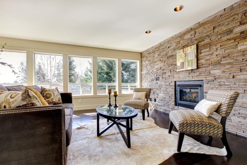 Living room with ledger stone fireplace wall, cushioned chairs, carpet floors, glass coffee table, and windows
