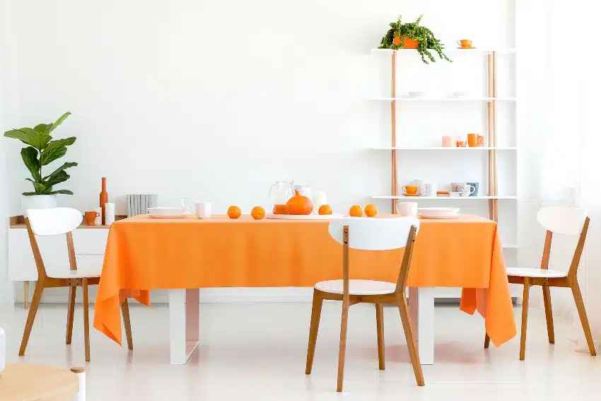 Long dining room table covered with orange tablecloth and comfortable white chairs around it