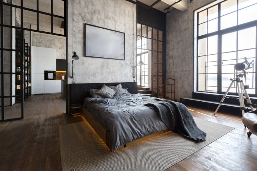 Modern industrial bedroom with wood floor, windows, concrete wall, lamp, comforter, and pillows