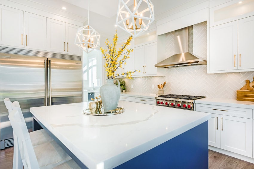 Modern kitchen with herringbone tile backsplash, white cabinets, countertop, island, flower vase, chairs, refrigerator, and pendant lights