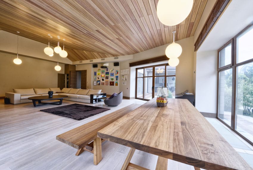 Modern living room with dining space, wood table, bench, couch, pendant lights, windows, wooden ceiling, and floors