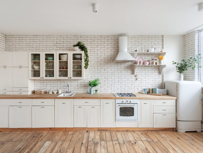 Modern one wall fitted kitchen facade at home with white interior, refrigerator, gas stove, cooking hood, built in oven, sink water tap, kitchenware supplies on shelves and plants on cupboard