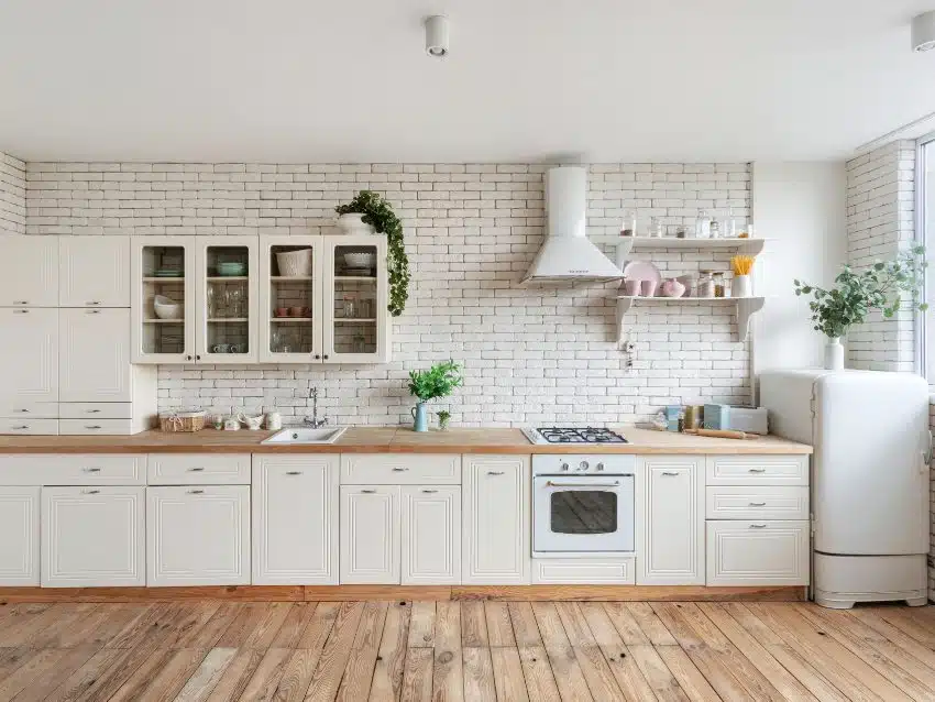 Modern one wall fitted kitchen facade at home with white interior, refrigerator, gas stove, cooking hood, built in oven, sink water tap, kitchenware supplies on shelves and plants on cupboard