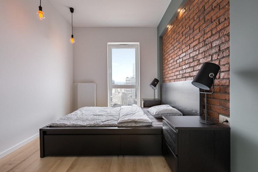 Rustic industrial bedroom with brick accent wall, wood flooring, nightstand, pillows, and window