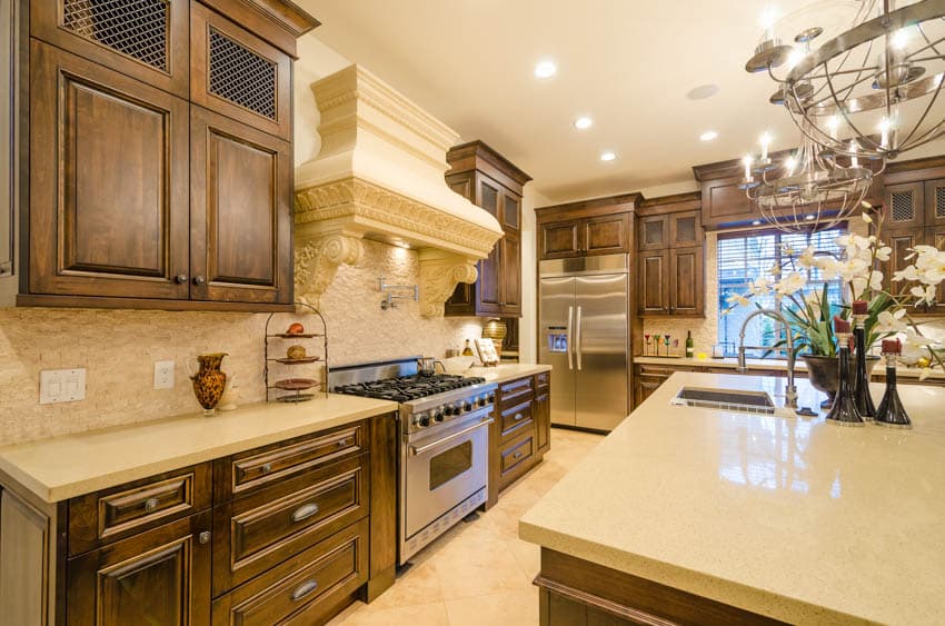 Rustic kitchen with wood cabinets, travertine backsplash, range hood, countertops, island, stove, oven, chandelier, and ceiling lights